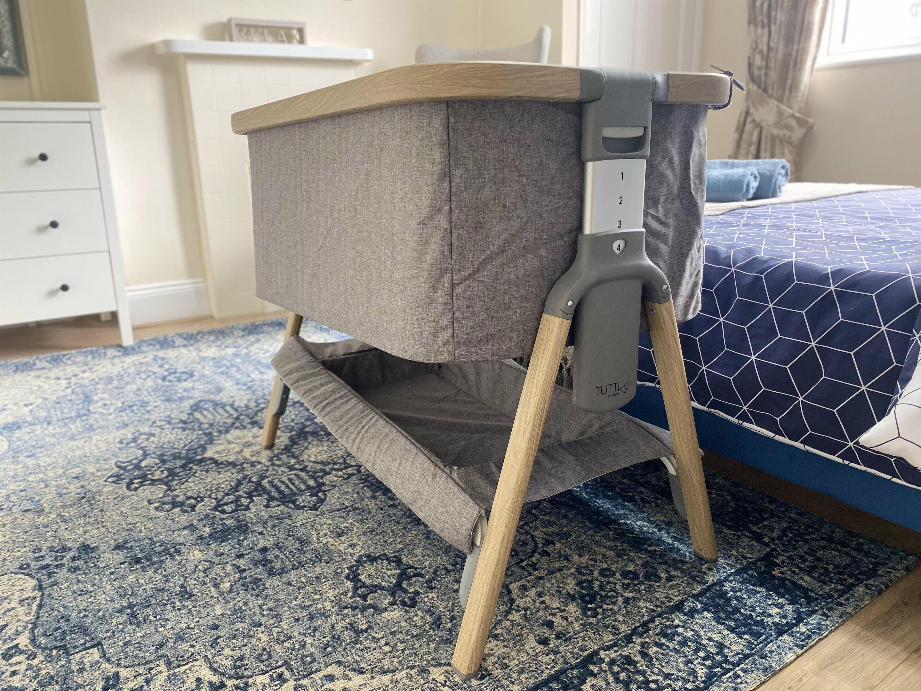 A baby rocker with a grey fabric seat and wooden legs, placed on a patterned rug in a room with a blue bed and a white dresser.