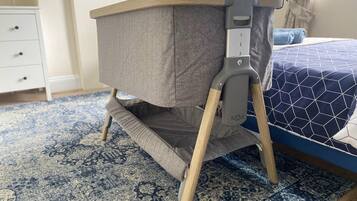 A baby rocker with a grey fabric seat and wooden legs, placed on a patterned rug in a room with a blue bed and a white dresser.