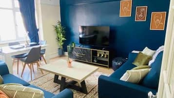 A living room with dark blue walls, a flat-screen TV, a sofa, a coffee table, and a dining area with a table and chairs.