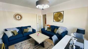 A living room with two blue sofas, a coffee table, a side table with a vase, and a door leading to another room.