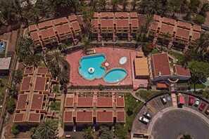 Ein Luftbild eines Hotels mit einem zentralen Schwimmbecken, mehreren Balkonen und umliegenden Palmen.