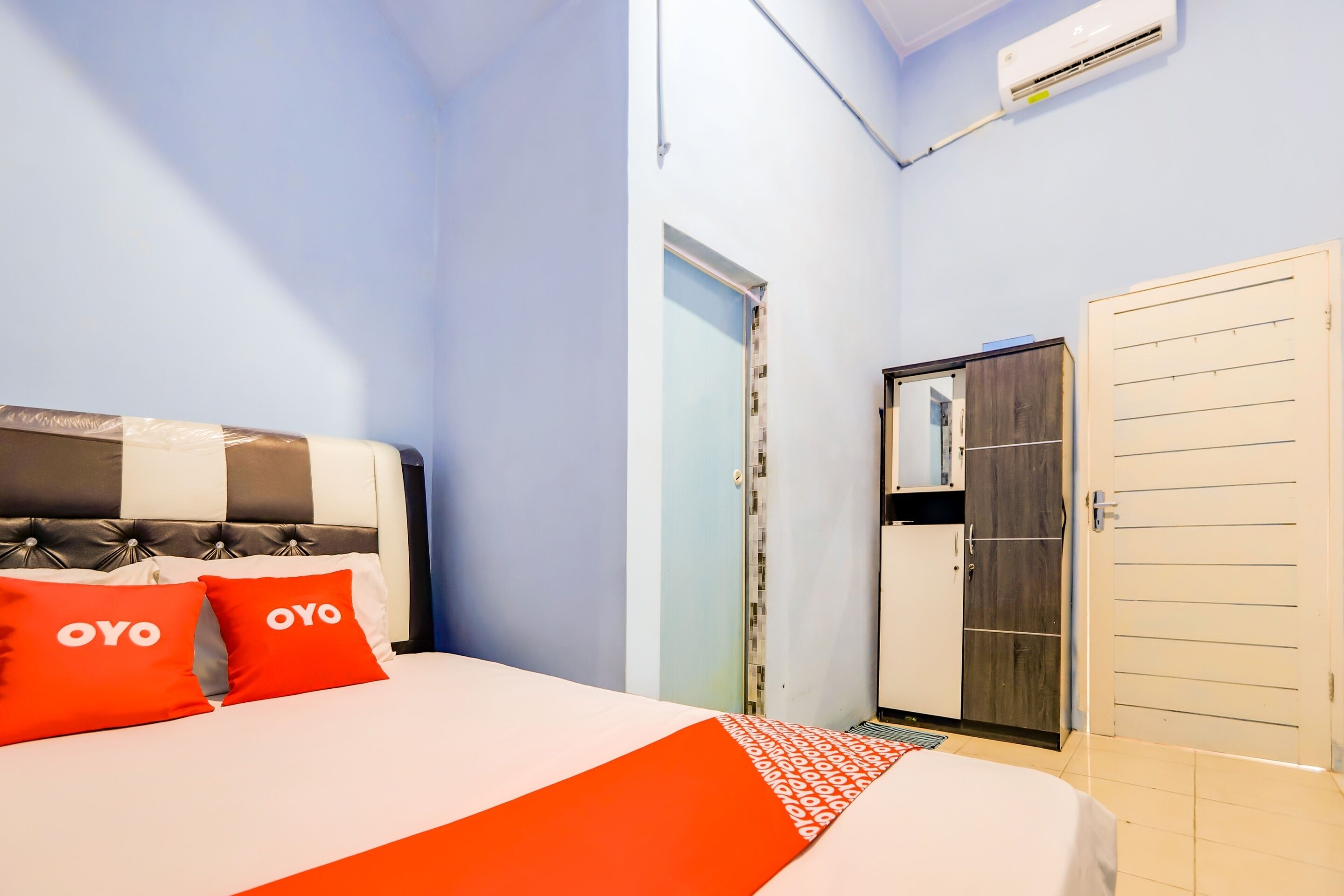 A bedroom with a bed, two red pillows with "OYO" text, a wardrobe, and an air conditioning unit.