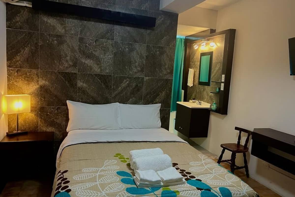 A hotel room with a bed, a nightstand, a bedside lamp, a bathroom with a sink and mirror, and a chair.