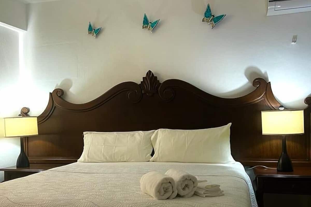 A neatly made bed with a patterned bedspread, two pillows, and rolled towels on the bed. There are two bedside lamps on either side of the bed. The room has a wooden headboard and a wall-mounted air conditioner.