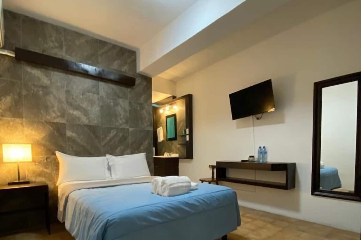 A modern hotel room with a large bed, a flat-screen TV, a stone accent wall, and a tiled floor.