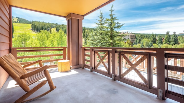 Ein hölzerner Schaukelstuhl auf einem Balkon mit Blick auf einen Wald und Gebäude in der Ferne.