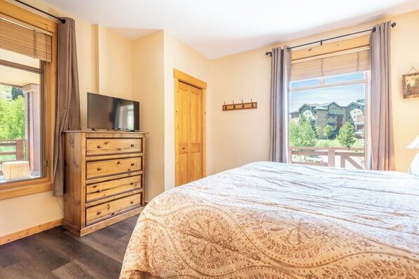A bedroom with a bed, a wooden dresser with a TV, a window with curtains, and a view of buildings outside.