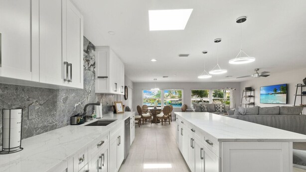 A modern kitchen with white cabinetry, a marble backsplash, and a central island. There is a dining area with a table and chairs, and a large window offering a view of a pool and greenery. - Serene Bayfront Stay - Dock, Hot Tub, Close to Beach (Bonita Springs)