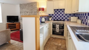A compact kitchen with white cabinets, a stove, a sink, and a television in a living area with a red rug and a stone accent wall. - Ty Bach Twt (Conwy)