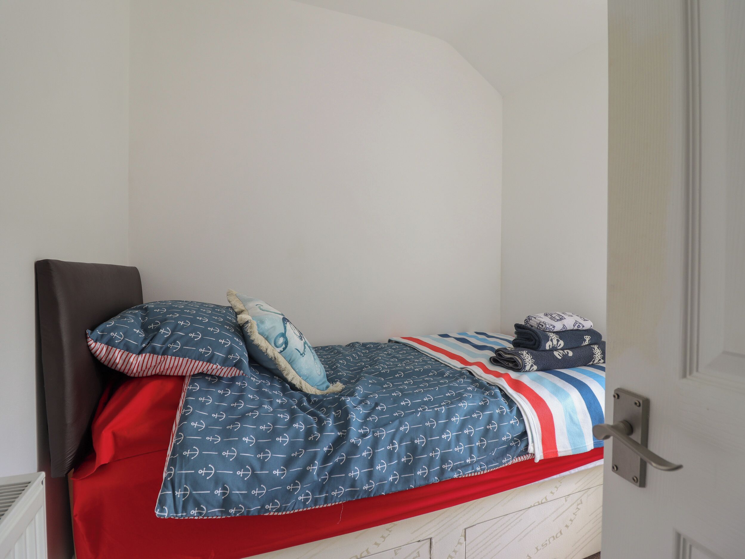 Una cama individual con una colcha a rayas azules y rojas, un almohadón con motivos náuticos y una toalla doblada al lado.