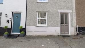 A two-story house with a white and grey textured exterior, a blue door, and a brown door. There are two windows, one with shutters and one without. - Ty Bach Twt (Conwy)