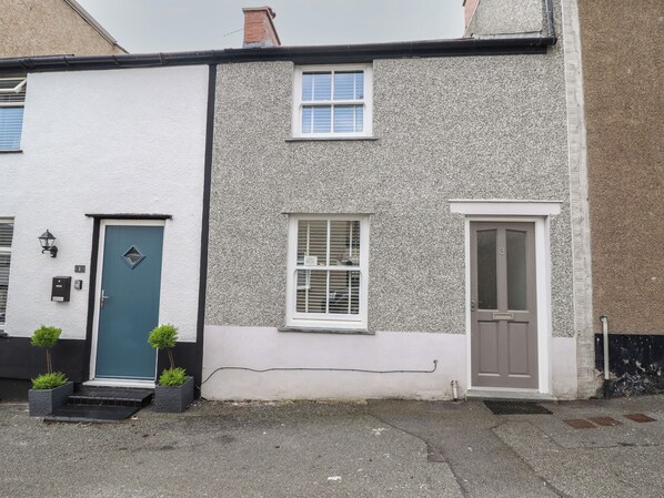 A two-story house with a white and grey textured exterior, a blue door, and a brown door. There are two windows, one with shutters and one without. - Ty Bach Twt (Conwy)