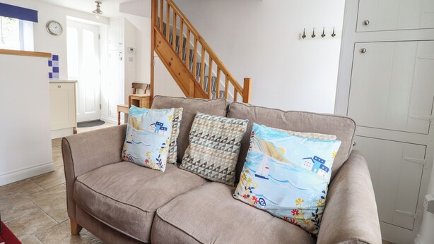 A living room with a beige sofa, patterned cushions, a wooden staircase, and a white door. - Ty Bach Twt (Conwy)