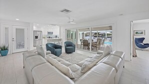 Un salon moderne avec un canapé blanc, deux fauteuils, une table basse et une grande fenêtre donnant sur un espace piscine.