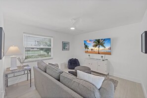 A living room with a sofa, a lamp, a ceiling fan, a window with blinds, a framed picture, and a wall-mounted TV.