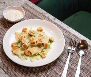 Une assiette de salade César, garnie de croûtons et de fromage râpé, un petit bol de vinaigrette, ainsi qu'une fourchette et une cuillère posées sur un set de table en fibres tressées.