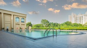 A swimming pool with a wooden deck, a building with arched windows, and a backdrop of high-rise buildings and greenery. - Apartel by Aarin - Oragadam (Sriperumbudur)