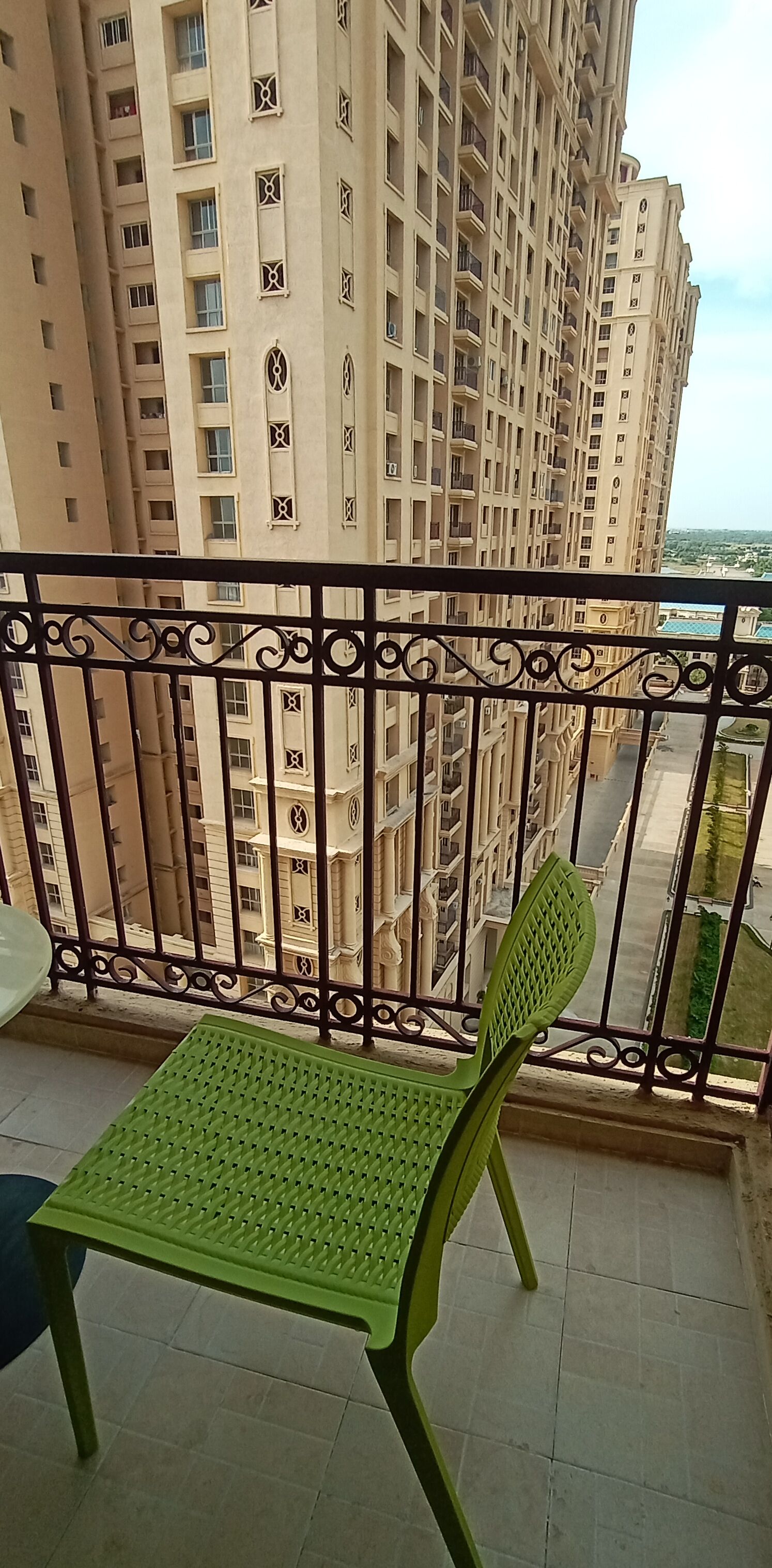 Uma varanda com uma cadeira verde, guarda-corpo de metal e vista para edifícios altos.