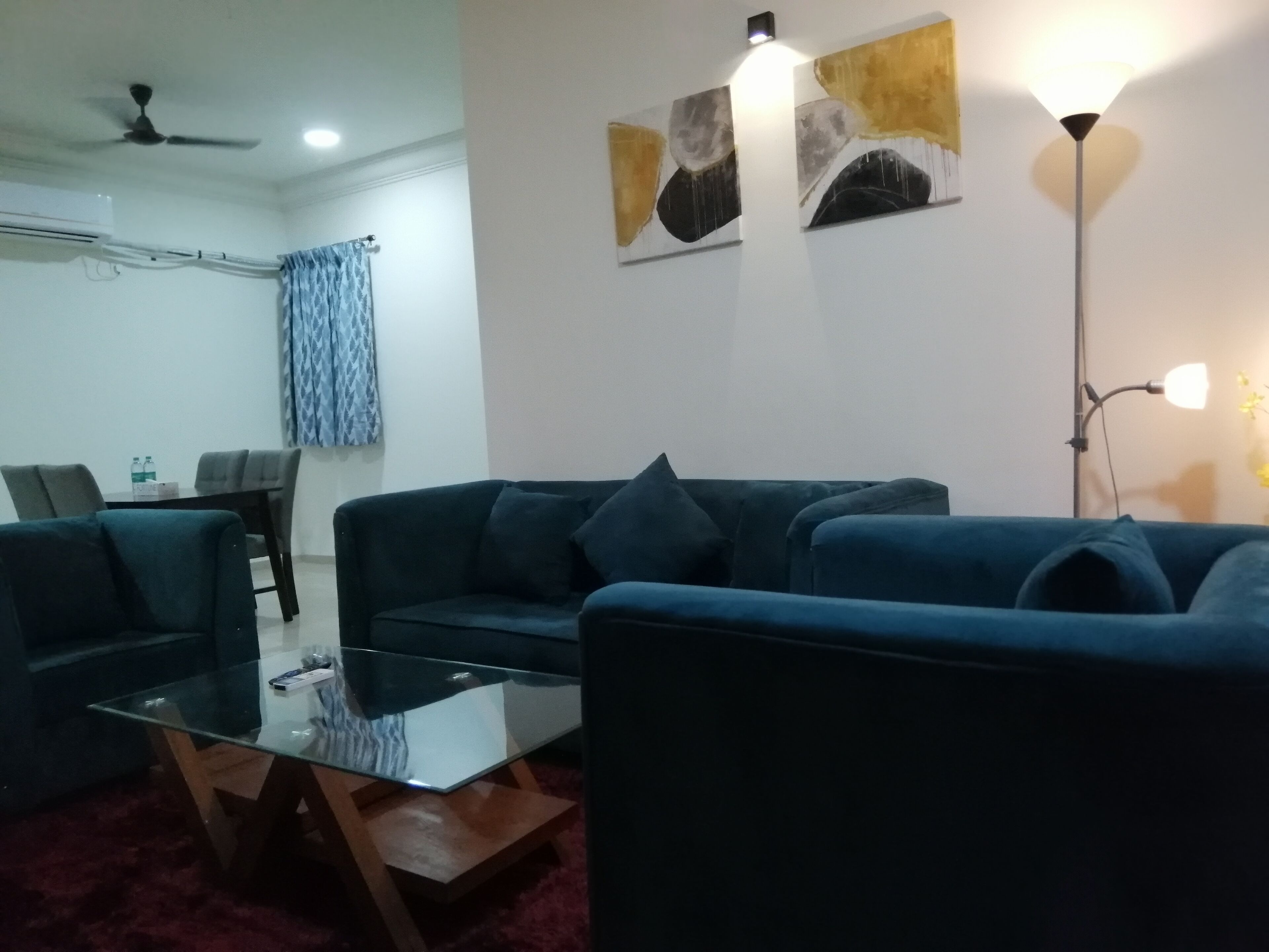 A living room with a dark blue sofa, a glass coffee table, and a ceiling fan. There are two wall-mounted lamps and abstract art on the wall.