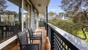 Een balkon met houten vloer en zwarte rieten stoelen, met uitzicht op een natuurlandschap met bomen en een heldere lucht.