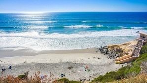 A beach with clear blue water, a sandy shore, and a building with a staircase leading down to the beach. - 10% OFF last minute stays - Oceanfront 1 bedroom, Pool, Spa, Beach Access (Solana Beach)