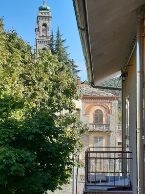 Ein Balkon mit einem dahinterliegenden Steinhaus, blauem Himmel und einem Baum im Vordergrund.