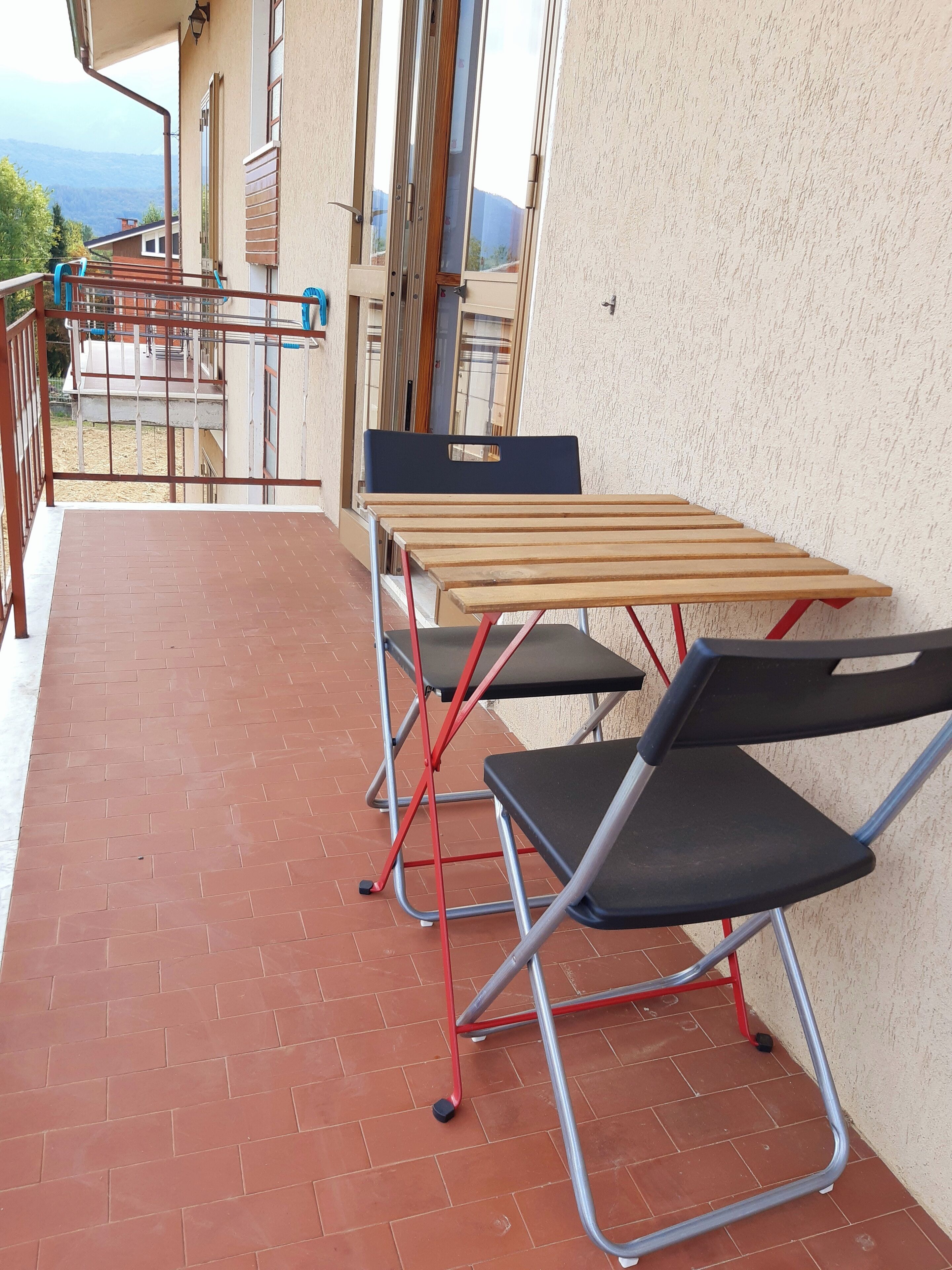 Un balcón con una mesa de madera y dos sillas plegables, vista a las montañas y un edificio con pared texturizada.