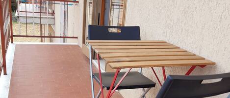 Un balcón con una mesa de madera y dos sillas plegables, vista a las montañas y un edificio con pared texturizada.