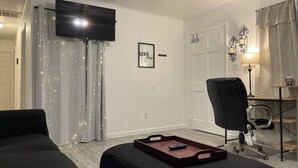 A living room with a black sofa, a black ottoman, a desk with a lamp, and a TV mounted on the wall. - Peaceful Retreat Near the Waterfront (Elizabeth City)