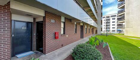 Un edificio moderno con fachada de ladrillo, una puerta negra con la etiqueta '103' y un pequeño jardín con arbustos y mantillo.