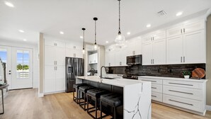 A modern kitchen with white cabinetry, a central island with stools, stainless steel appliances, and a black backsplash.