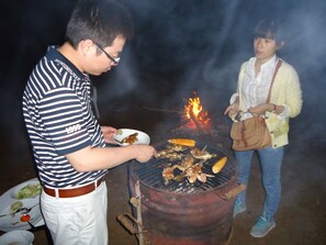 Una persona preparando comida a la parrilla al aire libre, con un fuego a la vista al fondo.
