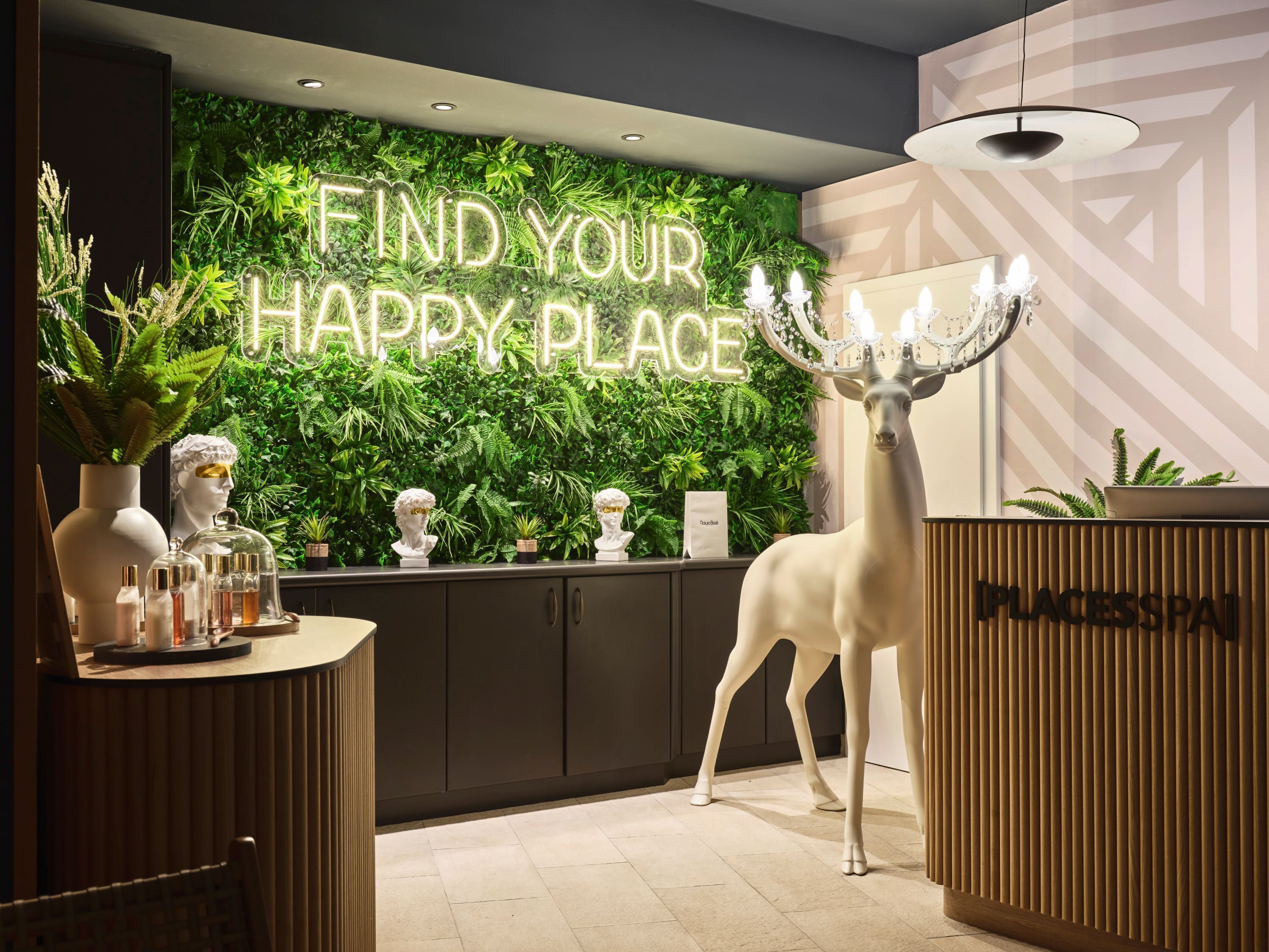 A reception area with a green wall, a neon sign reading "FIND YOUR HAPPY PLACE", a white deer statue, and a reception desk labeled "PLACESPAI".