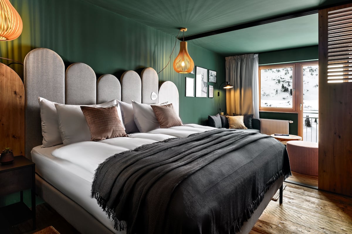 A bedroom with a large bed, a wooden headboard, a hanging light fixture, and a view of a snowy landscape through a window.