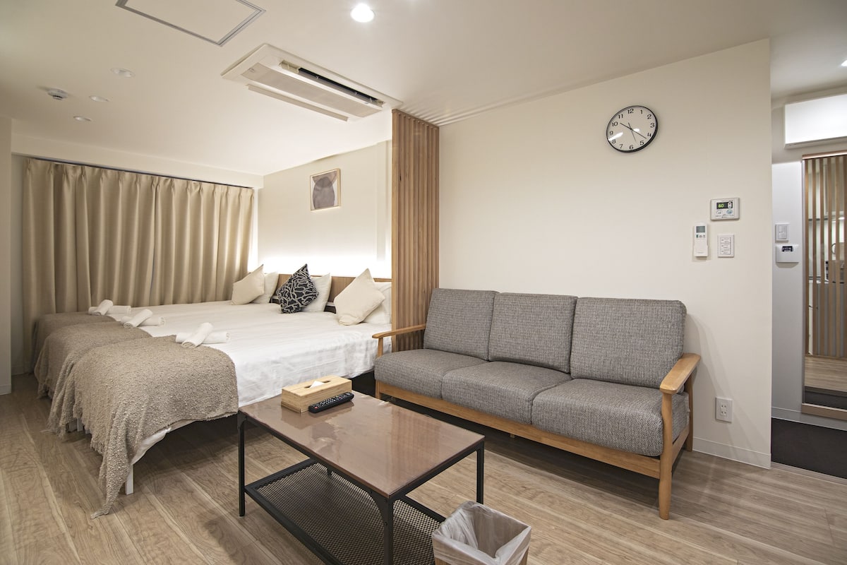 A hotel room with a bed, a grey sofa, a wooden coffee table, and a wall clock.