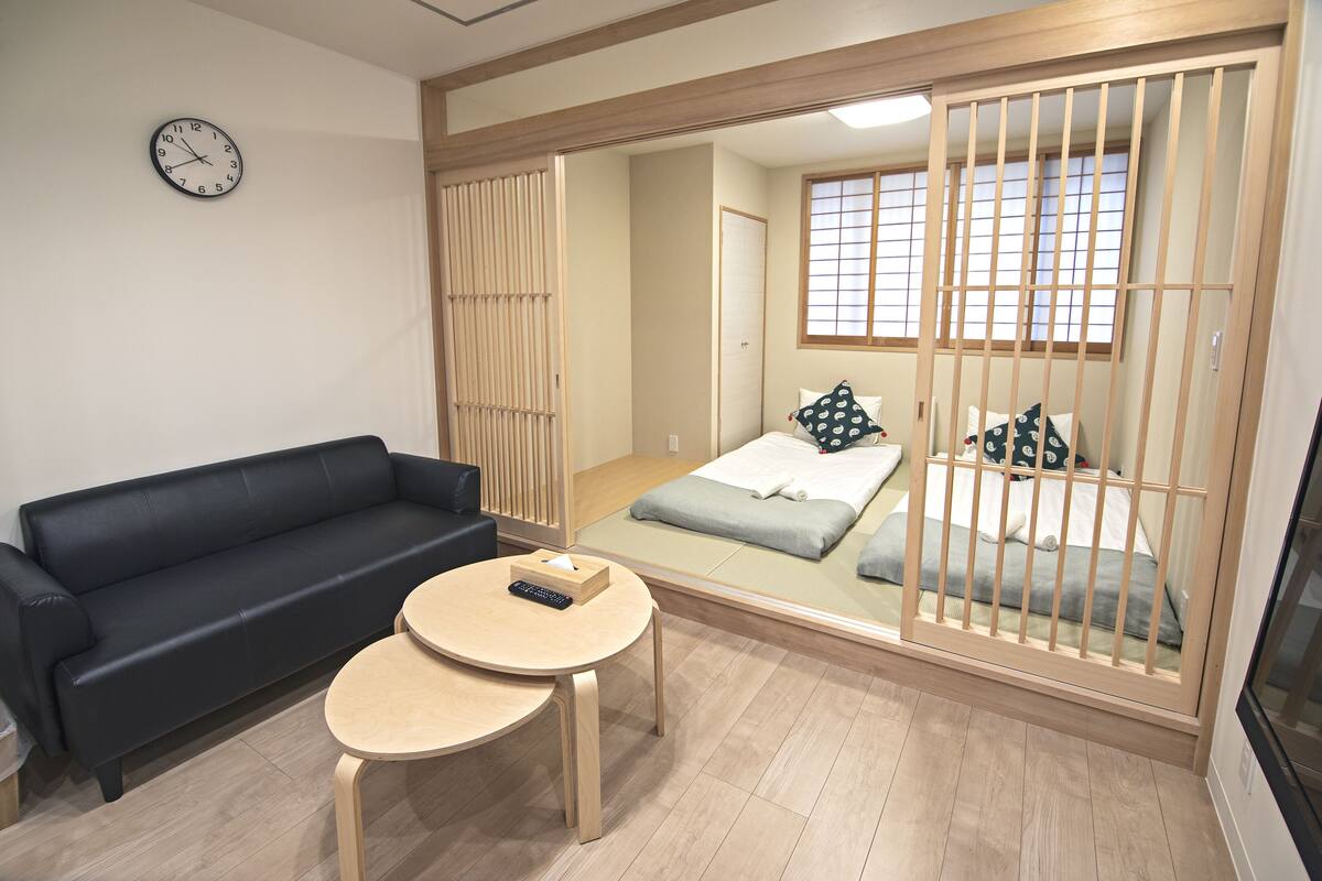A room with a black sofa, a small round table, a bed with a wooden frame, and a large mirror reflecting the room.