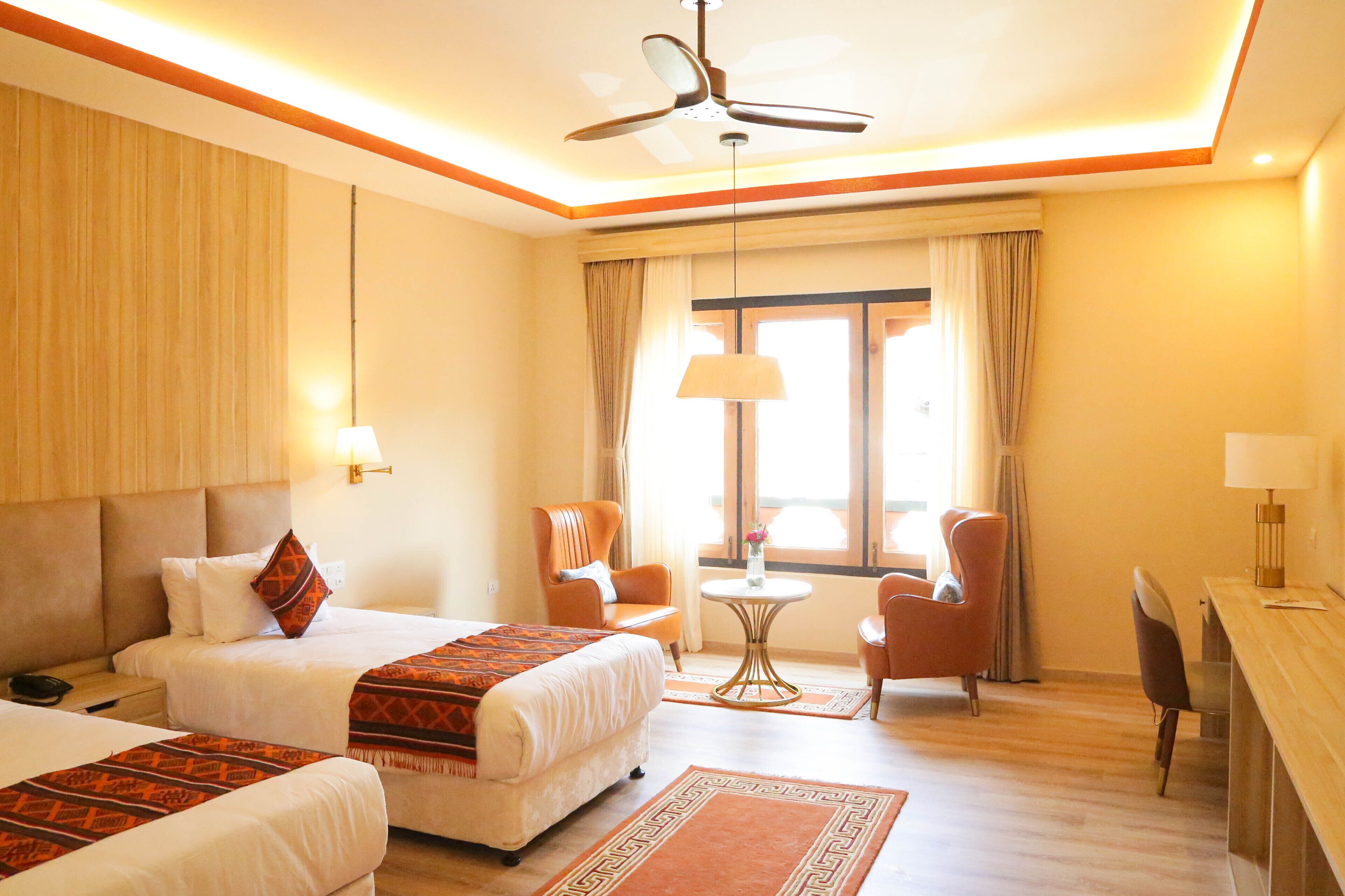 A hotel room with two beds, a desk, a chair, a ceiling fan, and a window with curtains.