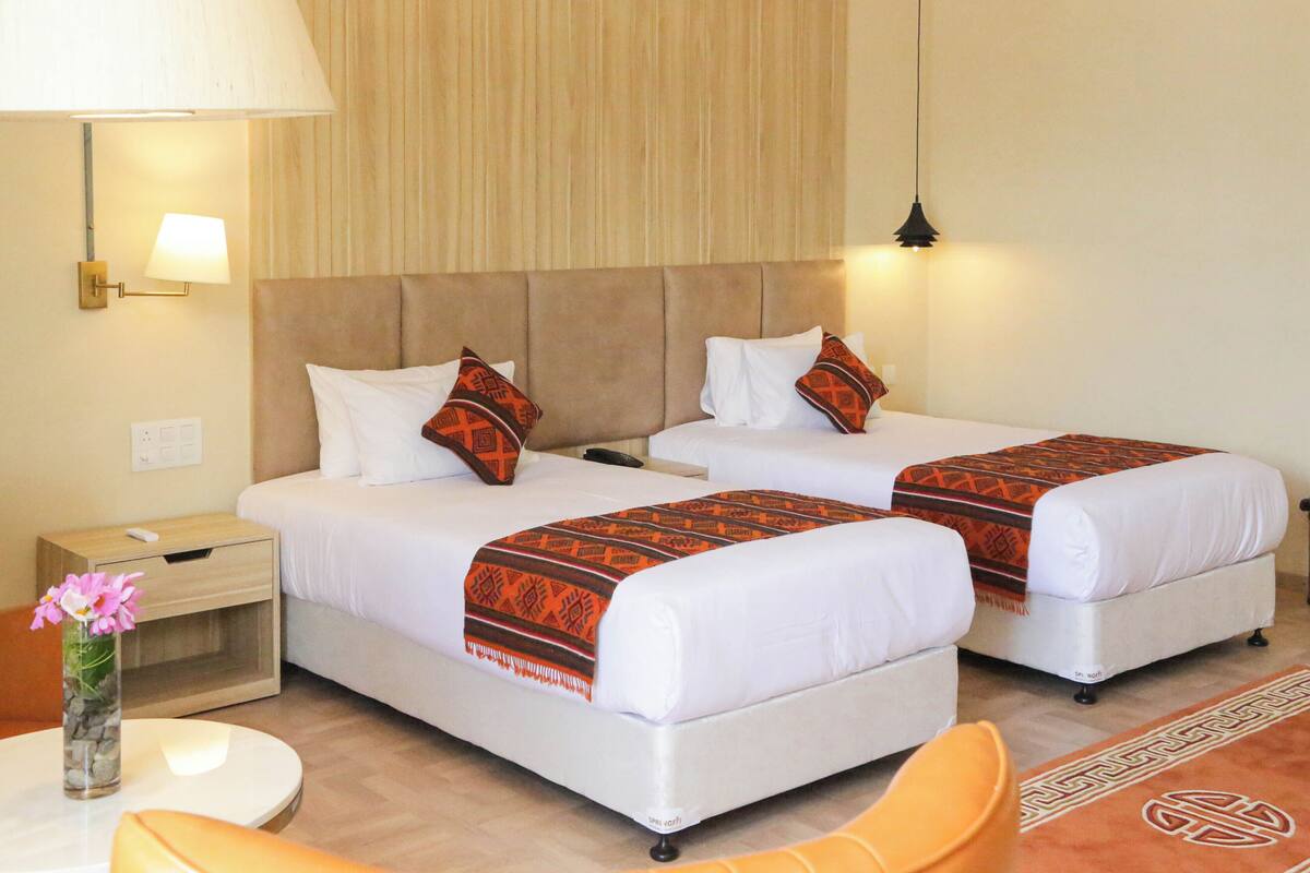 A hotel room with two beds, a nightstand, a lamp, a vase with flowers, and a chair.