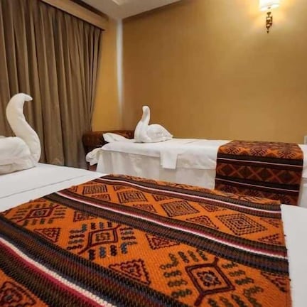 A hotel room with two beds, each with a white duvet and a decorative swan pillow. The room has a beige wall, a large window with curtains, and a wall-mounted light fixture.