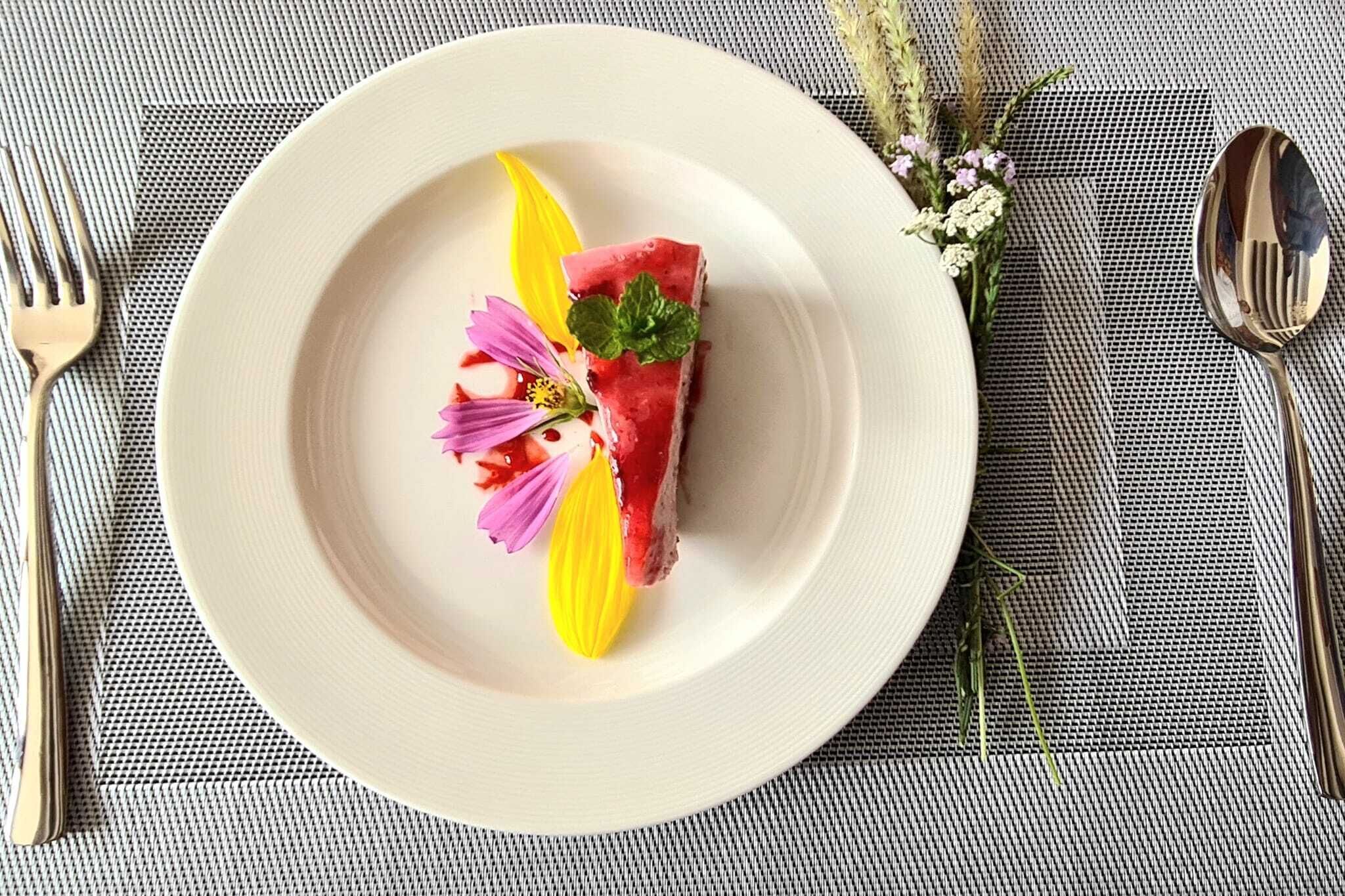 Ein Teller mit einer Wassermelone, einer gelben Frucht und violetten Blüten auf einer strukturierten Tischdecke.