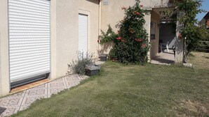 A building with a closed garage door, a small garden with grass and a flowering bush, and a tiled patio area. - House between sea, pool and garrigue (Cuxac-d'Aude)