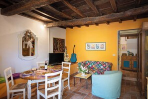 A dining area with a wooden table, chairs, and a television. There is a guitar and a colorful sofa in the background.