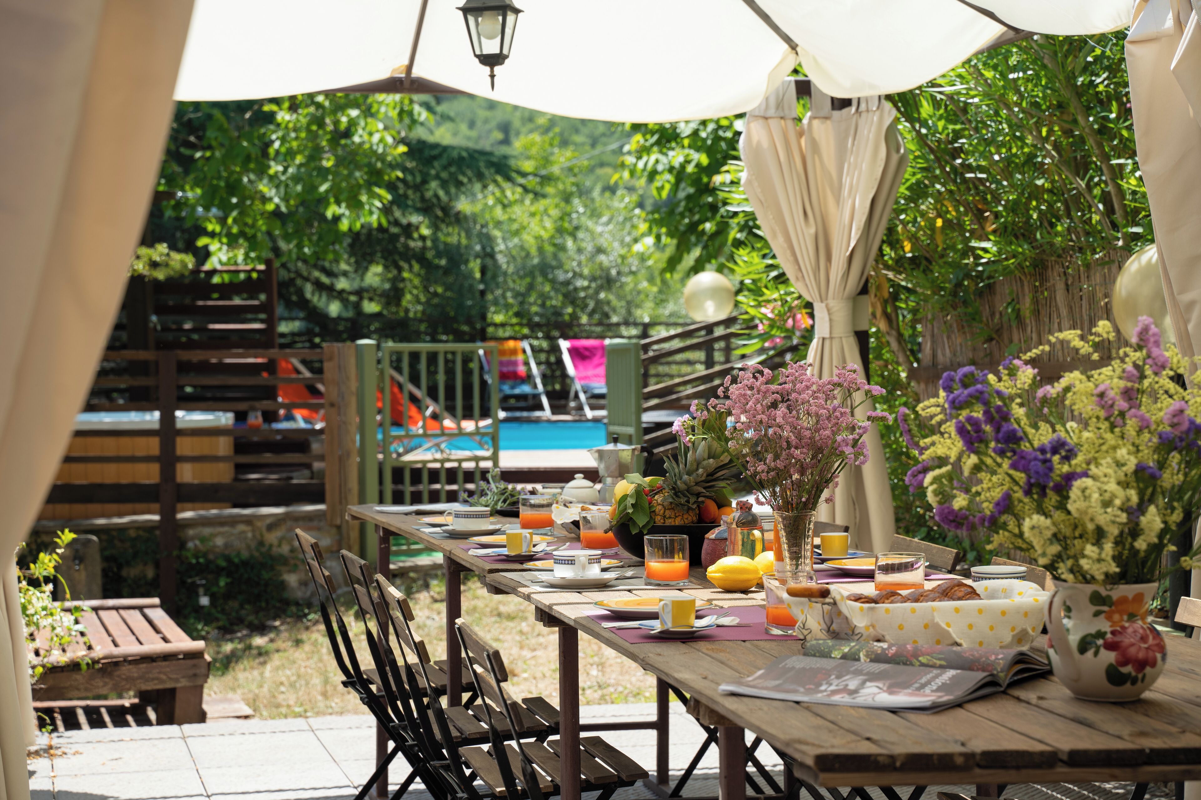 Un área de comedor al aire libre con una mesa de madera dispuesta para el desayuno, rodeada de sillas y con vistas a una piscina y follaje.