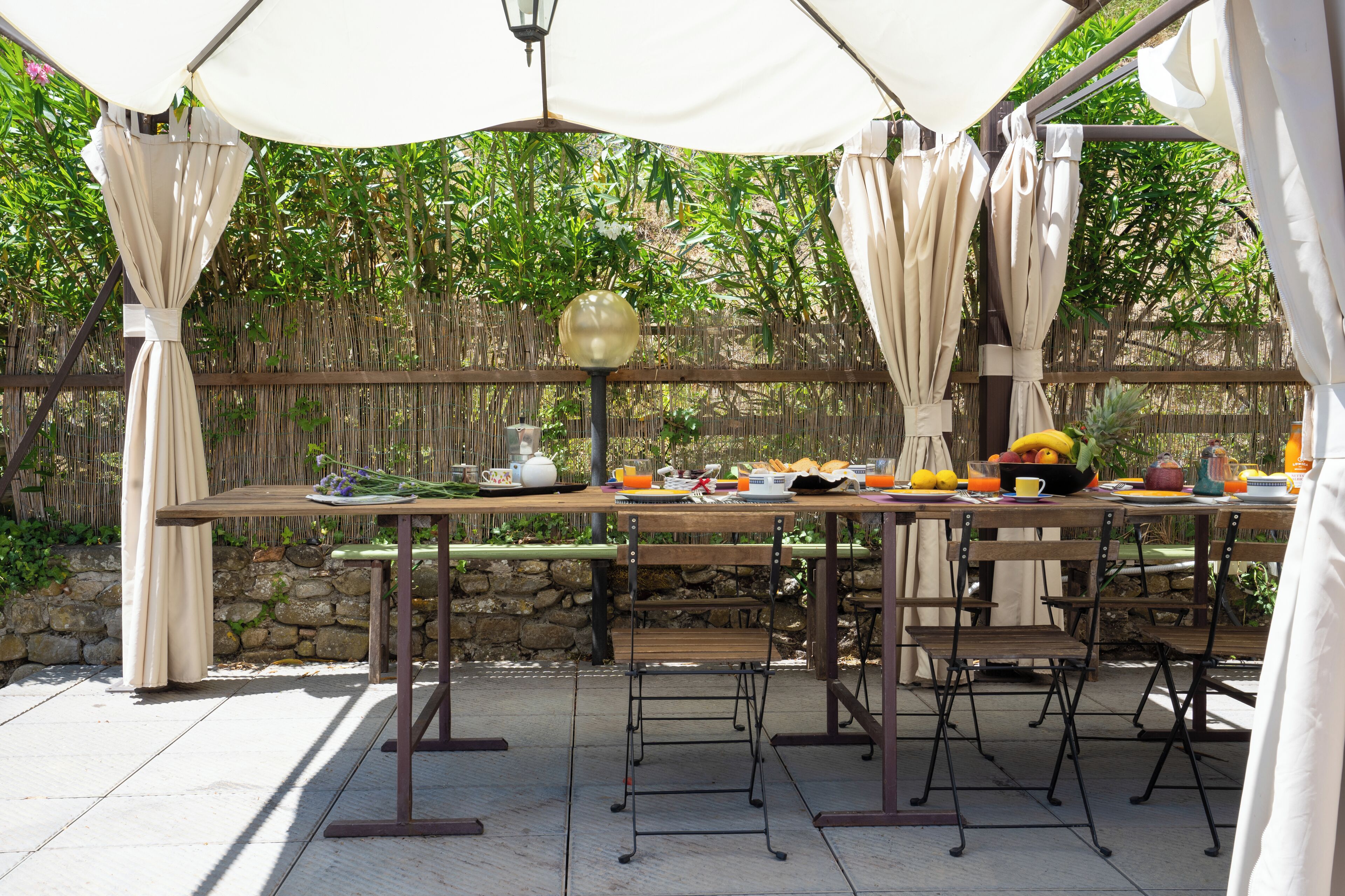 An outdoor dining area with a wooden table and metal chairs, surrounded by a bamboo fence and a stone wall.