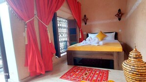 A bedroom with a bed, a red curtain, a window with metal bars, a decorative vase, and a red chair. - Riad La Bigarade (Marrakech)