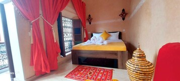 Un dormitorio con una cama, una cortina roja, una ventana con barrotes, un jarrón decorativo y una silla roja.