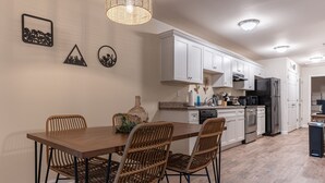 A modern kitchen with white cabinets, a wooden dining table, wicker chairs, and a hanging light fixture. - Near Atrium & town, King/Queen/Pullout, NO stairs (Lincolnton)