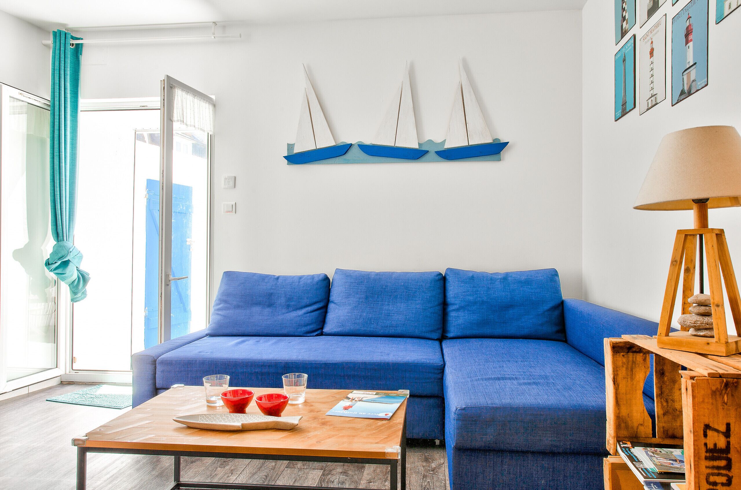 Ein Wohnzimmer mit einem blauen Sofa, einem Holztisch mit roten Schalen und Gläsern, einer Holzlampe und Wandkunst mit Segelbooten.