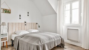 A bedroom with a sloped ceiling, a bed with a striped bedspread, a bedside table with a lamp, and a window with curtains. - Near the port of Guilvinec, town house for 5 (Le Guilvinec)