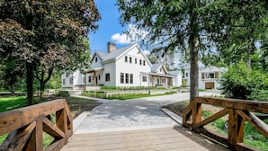 Un pont en bois mène à une maison blanche dotée d’une cheminée, entourée d’arbres et bordée d’un chemin pavé.